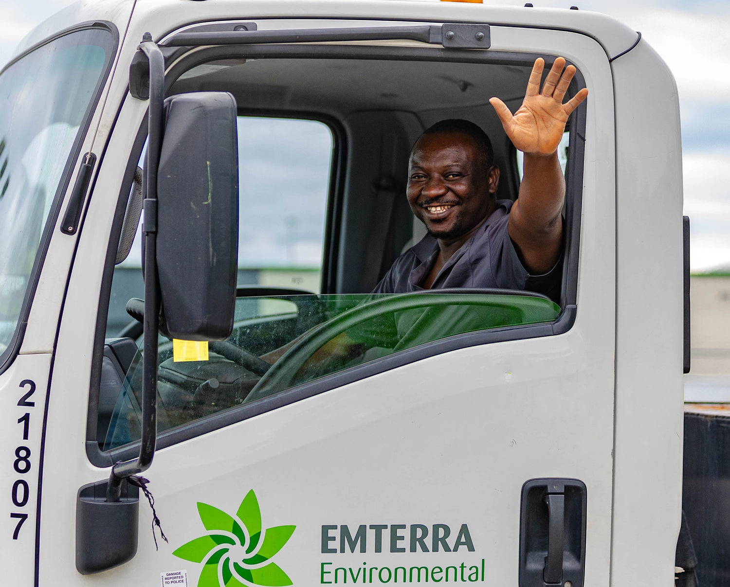 Person waving from inside an Emterra Environmental truck against a cloudy sky.